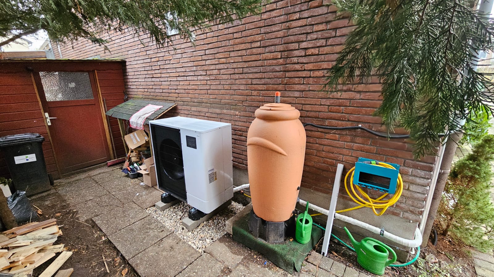 Außen aufgestellte Wärmepumpe neben Regenwassertank und Gartenschlauchbox an einer Backsteinwand unter Nadelbäumen.